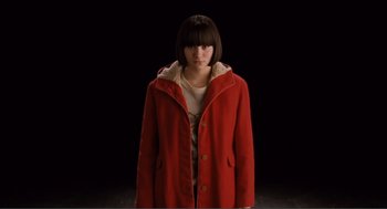 Movie still from “Submarine” (2010), directed by Richard Ayoade – A woman wearing a red coat standing in front of a black background; Medium shot, High angle