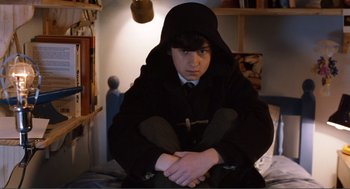 Movie still from “Submarine” (2010), directed by Richard Ayoade – A young man sitting on a bed wearing a hooded jacket; Medium shot, High angle