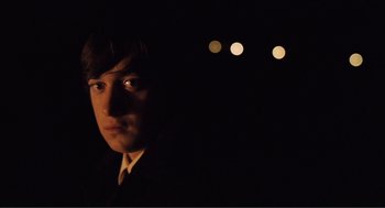 Movie still from “Submarine” (2010), directed by Richard Ayoade – A man in a suit and tie in the dark with lights in the background; Close Up shot, Over the shoulder angle