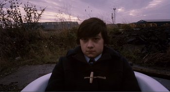 Movie still from “Submarine” (2010), directed by Richard Ayoade – A young man wearing a coat and tie with a cross on it; Medium shot, High angle