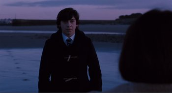 Movie still from “Submarine” (2010), directed by Richard Ayoade – A man in a black coat and tie standing in front of a body of water at night; Medium shot, Over the shoulder angle