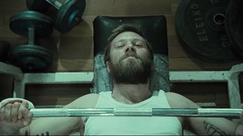 Movie still from “Submarino” (2010), directed by Thomas Vinterberg – A man with a beard is lifting a barbell in a gym; Medium shot, Low angle