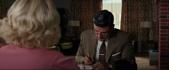 Movie still from “Suburbicon” (2017), directed by George Clooney – A man writing on a piece of paper while sitting at a table with an older woman; Medium shot, Over the shoulder angle