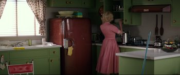 Movie still from “Suburbicon” (2017), directed by George Clooney – A woman in a pink dress in front of a green refrigerator; Wide shot, Over the shoulder angle
