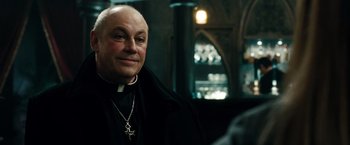 Movie still from “Sucker Punch” (2011), directed by Zack Snyder – A man in a priest's outfit smiling for the camera; Close Up shot, Over the shoulder angle