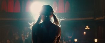 Movie still from “Sucker Punch” (2011), directed by Zack Snyder – A woman standing in front of a stage with a light shining on it; Medium shot, Over the shoulder angle