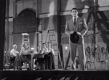 Movie still from “Sudden Fear” (1952), directed by David Miller – A black and white photo of a man in a suit holding a hat; Wide shot, Low angle