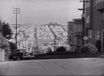 Movie still from “Sudden Fear” (1952), directed by David Miller – An old black and white photo of a car driving down a street; Extreme Wide shot, High angle