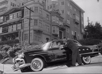 Movie still from “Sudden Fear” (1952), directed by David Miller – A black and white photo of a man leaning on a car; Wide shot, Low angle