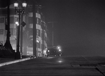 Movie still from “Sudden Fear” (1952), directed by David Miller – A car driving down a street at night in the fog; Extreme Wide shot, High angle