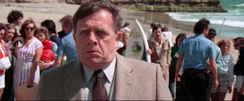 Movie still from “Sudden Impact” (1983), directed by Clint Eastwood – A man in a suit and tie standing in front of a group of people with surfboards; Close Up shot, Over the shoulder angle