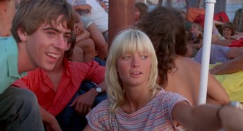 Movie still from “Summer Lovers” (1982), directed by Randal Kleiser – A woman with blonde hair sitting in front of a crowd; Close Up shot, Low angle