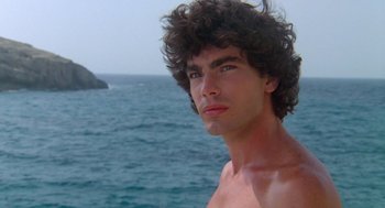 Movie still from “Summer Lovers” (1982), directed by Randal Kleiser – A young man with curly brown hair stands in front of a body of water; Close Up shot, Low angle
