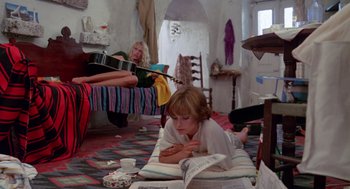 Movie still from “Summer Lovers” (1982), directed by Randal Kleiser – A woman reading a magazine while another woman plays a guitar; Wide shot, Low angle