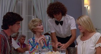 Movie still from “Summer Lovers” (1982), directed by Randal Kleiser – A woman sitting at a dinner table with a man standing next to her; Medium shot, Over the shoulder angle