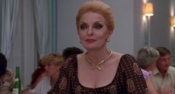 Movie still from “Summer Lovers” (1982), directed by Randal Kleiser – An older woman wearing a gold necklace and a brown dress; Close Up shot, Over the shoulder angle
