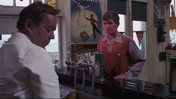 Movie still from “Summer of '42” (1971), directed by Robert Mulligan – A man standing at a counter in front of a bar; Medium shot, Over the shoulder angle