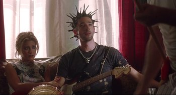 Movie still from “Summer of Sam” (1999), directed by Spike Lee – A man sitting on a couch playing a guitar; Medium shot, Over the shoulder angle