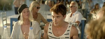 Movie still from “Suntan” (2016), directed by Argyris Papadimitropoulos – A young man in a striped tank top is sitting in front of a group of people; Medium shot, Over the shoulder angle