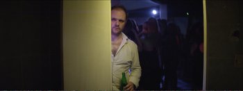 Movie still from “Suntan” (2016), directed by Argyris Papadimitropoulos – A man holding a beer standing in front of a wall; Medium shot, Over the shoulder angle