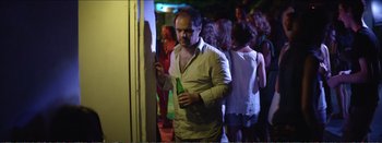 Movie still from “Suntan” (2016), directed by Argyris Papadimitropoulos – A man holding a beer in his hand while standing in front of a wall; Medium shot, Over the shoulder angle