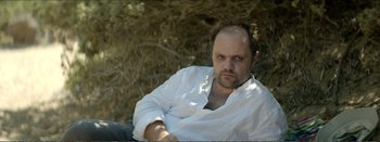 Movie still from “Suntan” (2016), directed by Argyris Papadimitropoulos – A man in a white shirt sitting in front of some trees; Close Up shot, Low angle