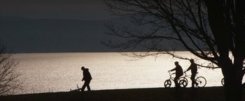 Movie still from “Super Dark Times” (2017), directed by Kevin Phillips – A man walking next to a bike on the shore of a body of water; Extreme Wide shot, Low angle