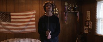 Movie still from “Super Dark Times” (2017), directed by Kevin Phillips – A man holding a sword in a room with trophies on the wall; Medium shot, Low angle