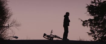 Movie still from “Super Dark Times” (2017), directed by Kevin Phillips – A man standing next to a bicycle on the side of the road; Wide shot, Low angle