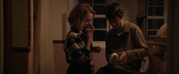 Movie still from “Super Dark Times” (2017), directed by Kevin Phillips – A woman and a man looking at a cell phone in a room; Medium shot, Over the shoulder angle