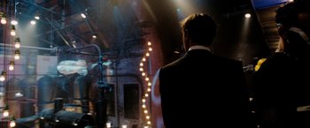 Movie still from “Surrogates” (2009), directed by Jonathan Mostow – A man standing in front of a stage with lights in the background; Wide shot, Over the shoulder angle