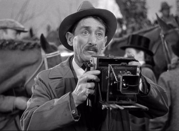Movie still from “Suspicion” (1941), directed by Alfred Hitchcock – An older man holding a camera in a crowd of people; Close Up shot, Low angle