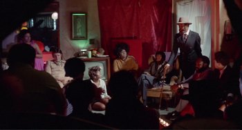 Movie still from “Sweet Sweetback's Baadasssss Song” (1971), directed by Melvin Van Peebles – A group of people sitting in a living room; Wide shot, Low angle