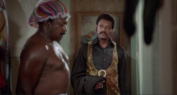 Movie still from “Sweet Sweetback's Baadasssss Song” (1971), directed by Melvin Van Peebles – A man in a black shirt is looking in the mirror while another man stands next to him; Medium shot, Over the shoulder angle