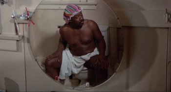 Movie still from “Sweet Sweetback's Baadasssss Song” (1971), directed by Melvin Van Peebles – A man sitting on a toilet in a bathroom; Medium shot, Low angle