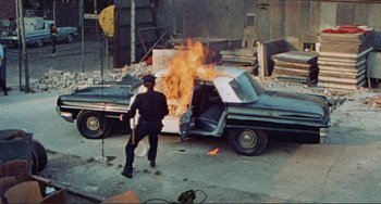 Movie still from “Sweet Sweetback's Baadasssss Song” (1971), directed by Melvin Van Peebles – A man standing in front of a car on fire; Wide shot, High angle