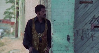 Movie still from “Sweet Sweetback's Baadasssss Song” (1971), directed by Melvin Van Peebles – A man wearing a gold vest standing next to a brick wall; Medium shot, Over the shoulder angle