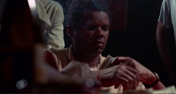 Movie still from “Sweet Sweetback's Baadasssss Song” (1971), directed by Melvin Van Peebles – A man sitting at a table with a cigarette in his hand; Close Up shot, Low angle