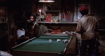 Movie still from “Sweet Sweetback's Baadasssss Song” (1971), directed by Melvin Van Peebles – A man sitting in front of a pool table in a room; Wide shot, High angle
