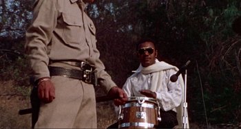 Movie still from “Sweet Sweetback's Baadasssss Song” (1971), directed by Melvin Van Peebles – A man playing a drum in front of another man in uniform; Medium shot, Low angle