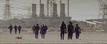 Movie still from “Syriana” (2005), directed by Stephen Gaghan – A group of people walking on a beach near a power plant; Wide shot, Low angle