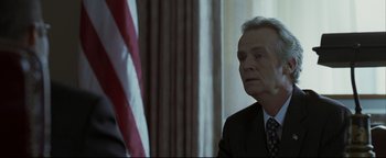 Movie still from “Syriana” (2005), directed by Stephen Gaghan – A man in a suit and tie in front of an american flag; Close Up shot, Low angle