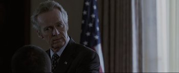 Movie still from “Syriana” (2005), directed by Stephen Gaghan – An older man wearing a suit and tie in front of an american flag; Close Up shot, Over the shoulder angle