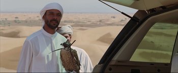 Movie still from “Syriana” (2005), directed by Stephen Gaghan – A man holding a bird in his hand while standing in front of a vehicle; Close Up shot, Over the shoulder angle