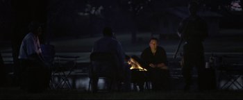 Movie still from “Syriana” (2005), directed by Stephen Gaghan – Two people sitting at a table in the dark with a fire; Wide shot, Over the shoulder angle