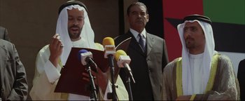 Movie still from “Syriana” (2005), directed by Stephen Gaghan – A man is speaking at a press conference; Medium shot, Over the shoulder angle