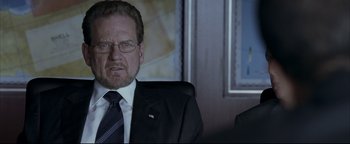 Movie still from “Syriana” (2005), directed by Stephen Gaghan – A man in a suit and tie sitting in a chair; Close Up shot, Over the shoulder angle