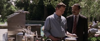 Movie still from “Syriana” (2005), directed by Stephen Gaghan – A man standing in front of a table with plates of food on top of it; Medium shot, Over the shoulder angle