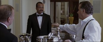 Movie still from “Syriana” (2005), directed by Stephen Gaghan – A man in a tuxedo is drinking from a glass; Medium shot, Over the shoulder angle
