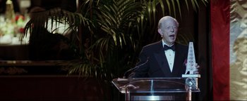 Movie still from “Syriana” (2005), directed by Stephen Gaghan – An older man in a tuxedo speaking at an event; Medium shot, High angle